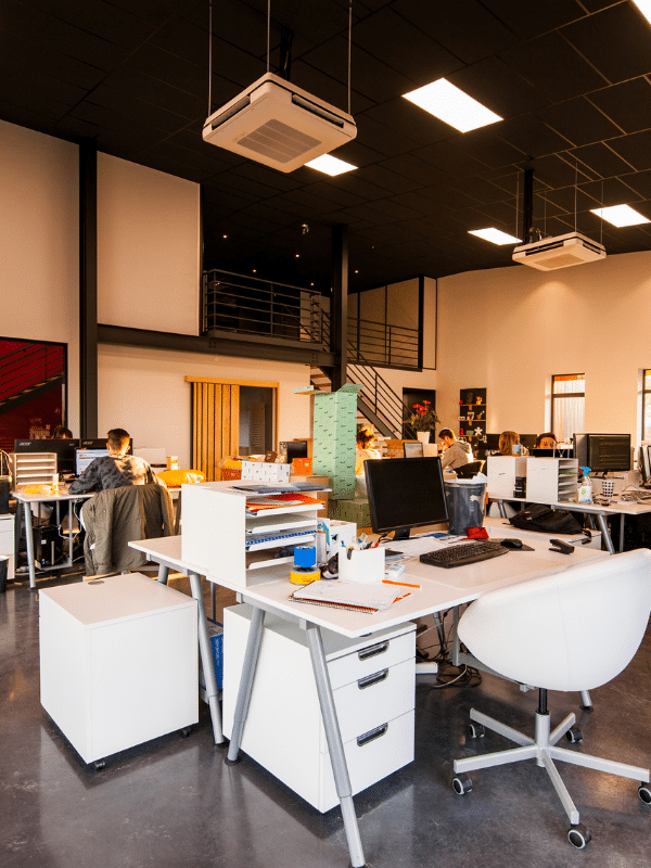Modern open-plan office space with several desks, computers, and office chairs. People are working at their stations. The office has high ceilings, large windows, and a mezzanine level in the background.