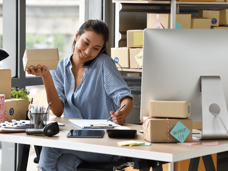 A woman sits at a desk in a modern office, talking on the phone and smiling while holding a small package. The desk is cluttered with parcels, stationery, and a computer, suggesting she is running an online business.