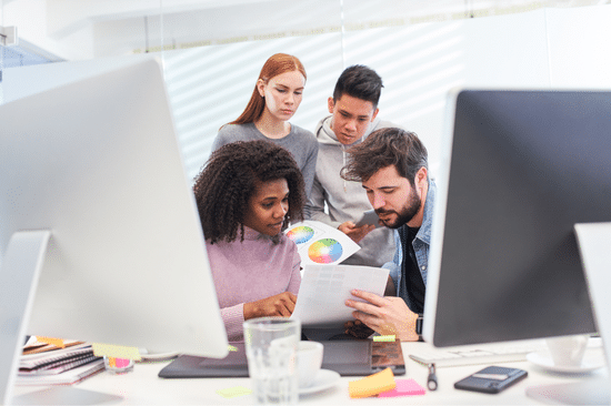 Four colleagues gather around a desk in a modern office, reviewing documents and a color wheel, with computer monitors and office supplies visible in the foreground.