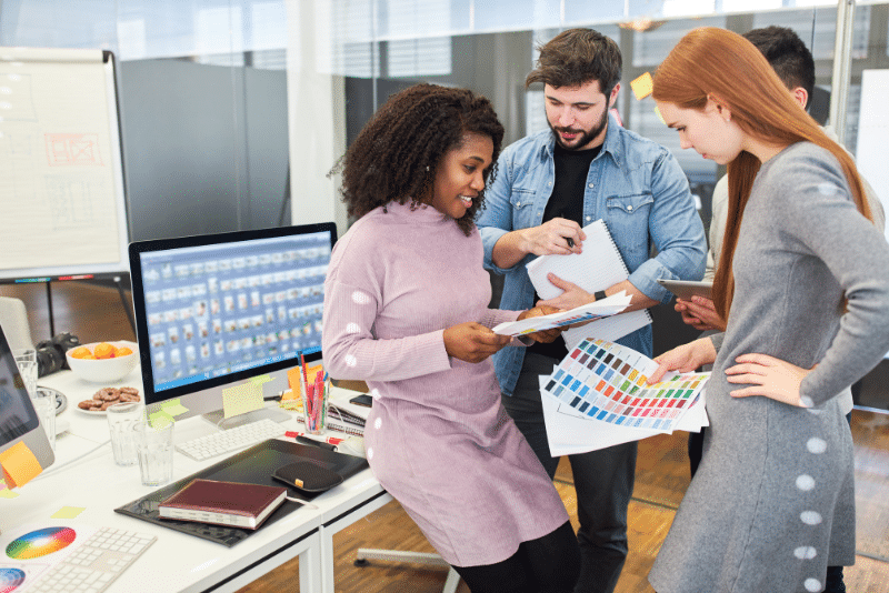 Four colleagues in a modern office discuss color swatches and notes, collaborating on a creative project. A computer monitor, office supplies, and charts are visible in the workspace.